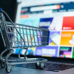 Ein Miniatur-Einkaufswagen steht auf der Tastatur eines Laptops, während auf dem Bildschirm eine E-Commerce-Website mit bunten Produktangeboten angezeigt wird. Die Szene symbolisiert Online-Shopping und digitale Einkaufsprozesse