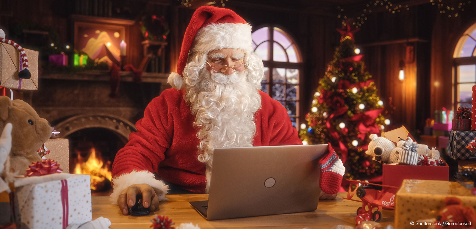 Weihnachtskommunikation Social Media: Weihnachtsmann am Laptop im Büro vor Kamin und Weihnachtsbaum, plant Social Media Postings.